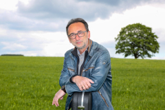 Lutz Scheufler gestützt auf Gitarrenkoffer vor grünem Feld mit Baum