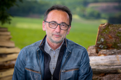 Lutz Scheufler (Nahaufnahme). Rechts und links Stapel mit gefällten Baumstämmen. Im Hintergrund Waldwiese