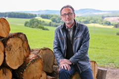 Lutz Scheufler neben Stapel mit gefällten Baumstämmen. Im Hintergrund Wald, Felder und Hügel