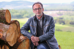 Lutz Scheufler neben Stapel mit gefällten Baumstämmen. Im Hintergrund Wald, Felder und Hügel